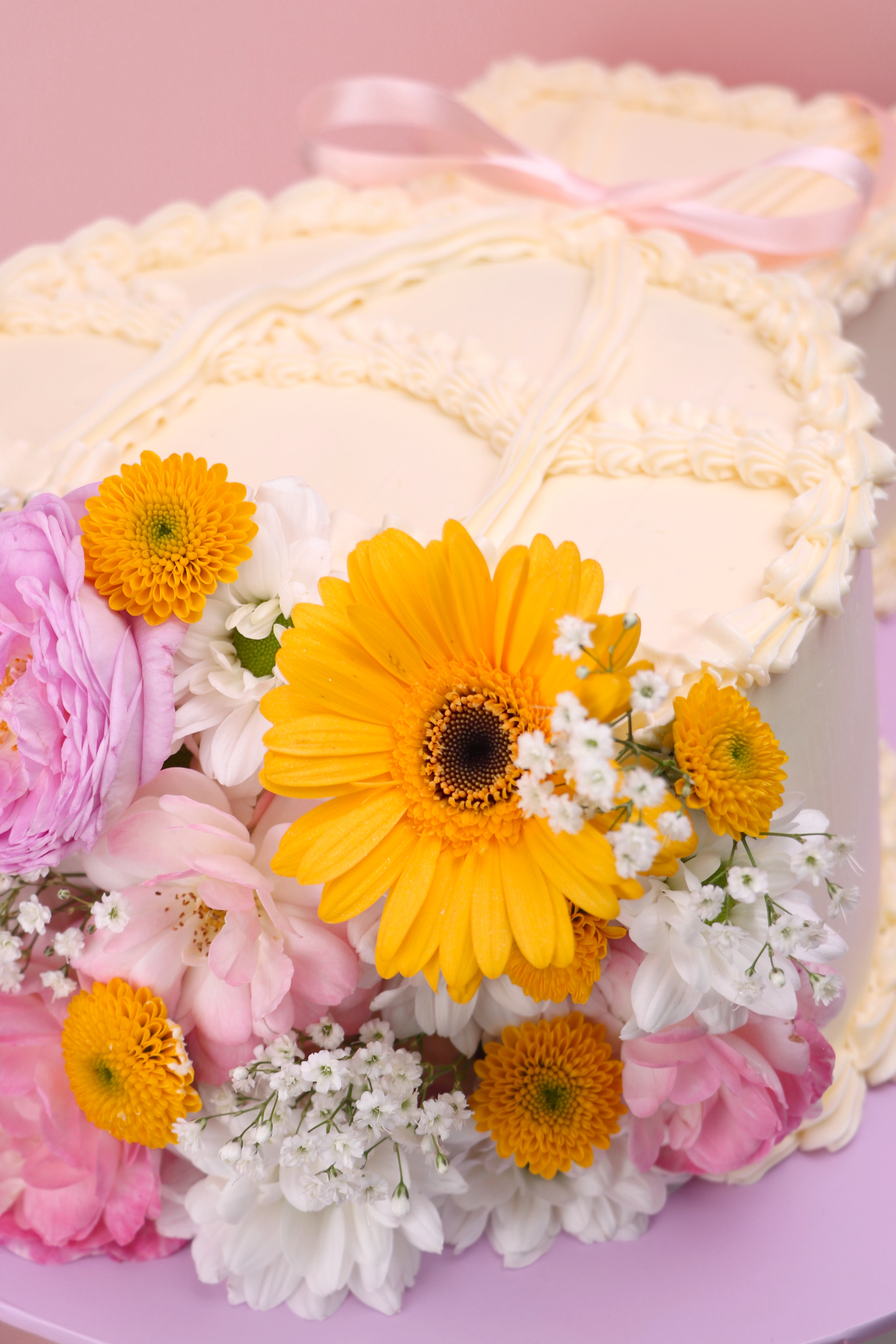 Heart-shaped fresh flower cake with yellow daisies, pink roses and vintage piped buttercream on lilac board handmade by Cakeasion in Uxbridge.