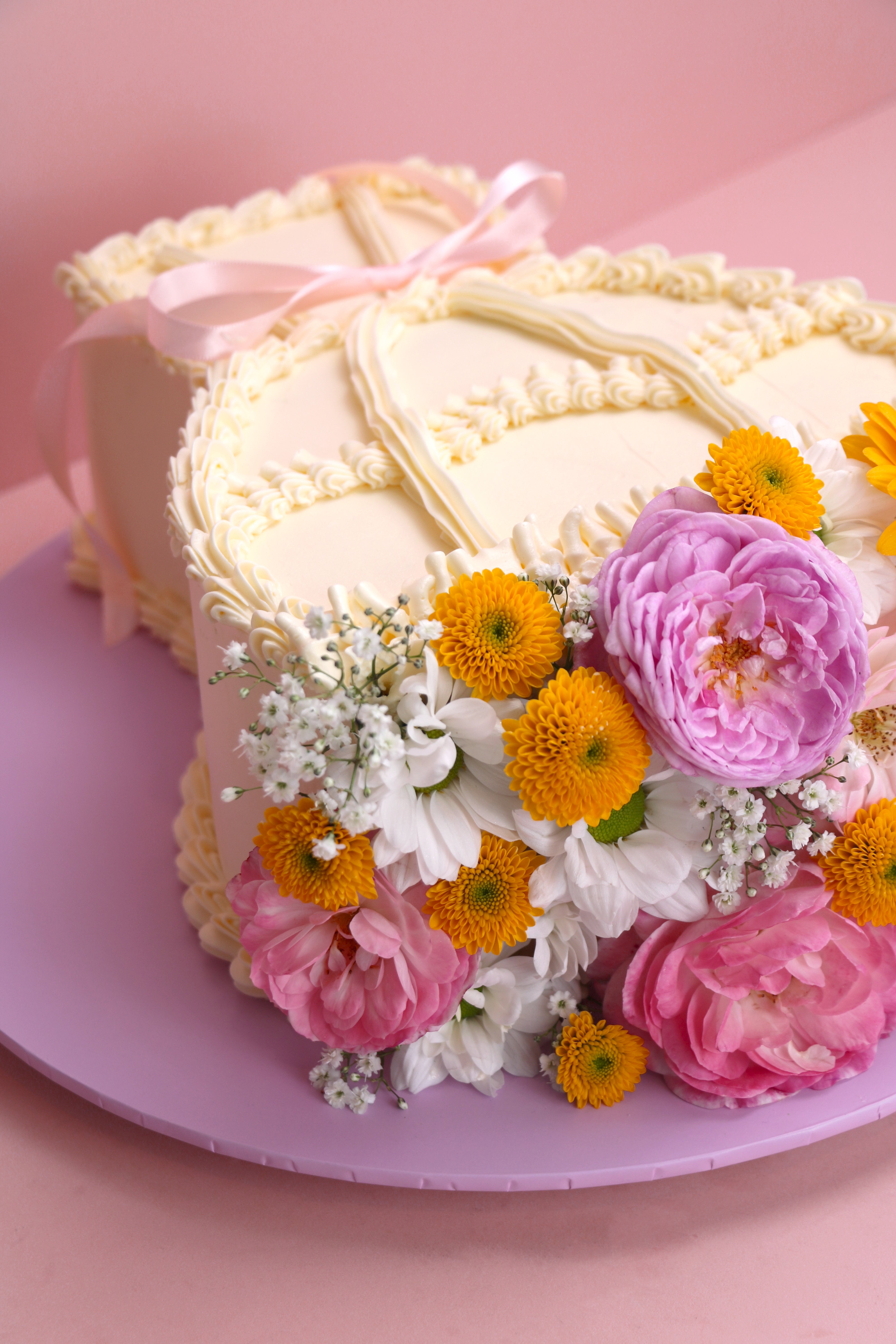 Heart-shaped fresh flower cake with yellow daisies, pink roses and vintage piped buttercream on lilac board handmade by Cakeasion in Uxbridge.