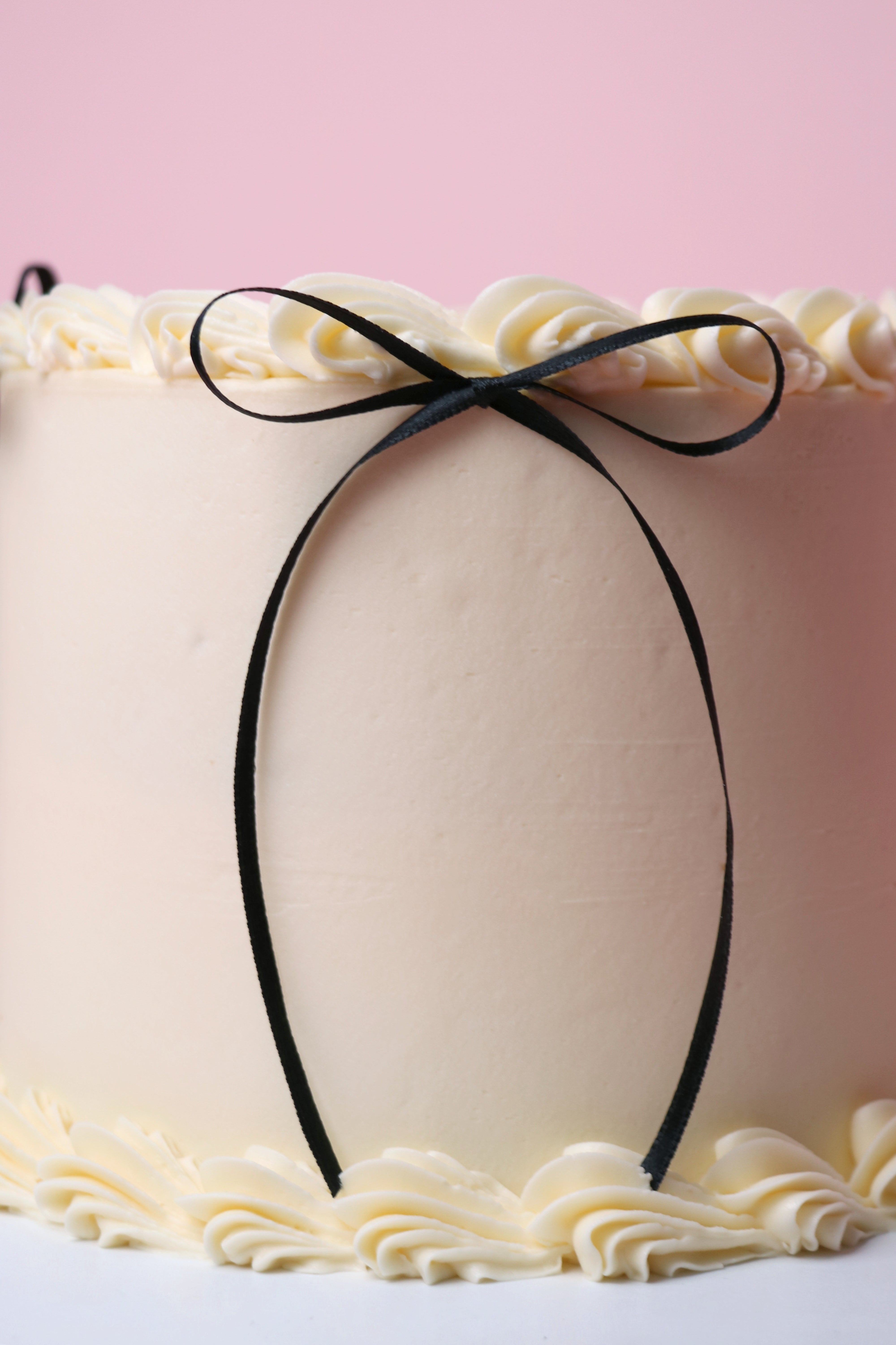 Close-up of a cake with white frosting and a black ribbon against a pink background. Elegant black and white celebration cake – perfect for birthdays, engagements, and events in West London.