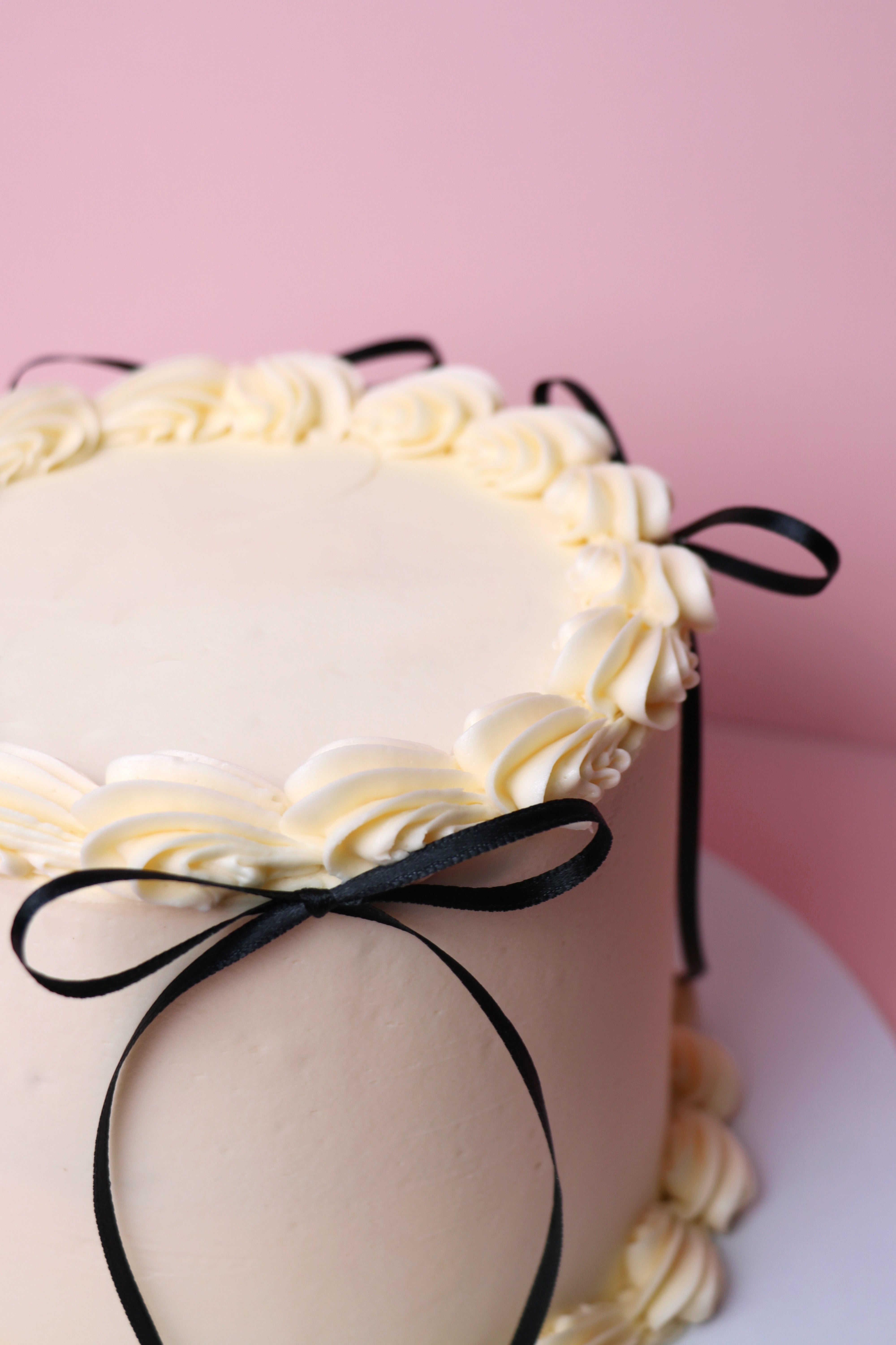 White cake with black ribbons on a pink background. Handmade in Uxbridge by Cakeasion Shop.