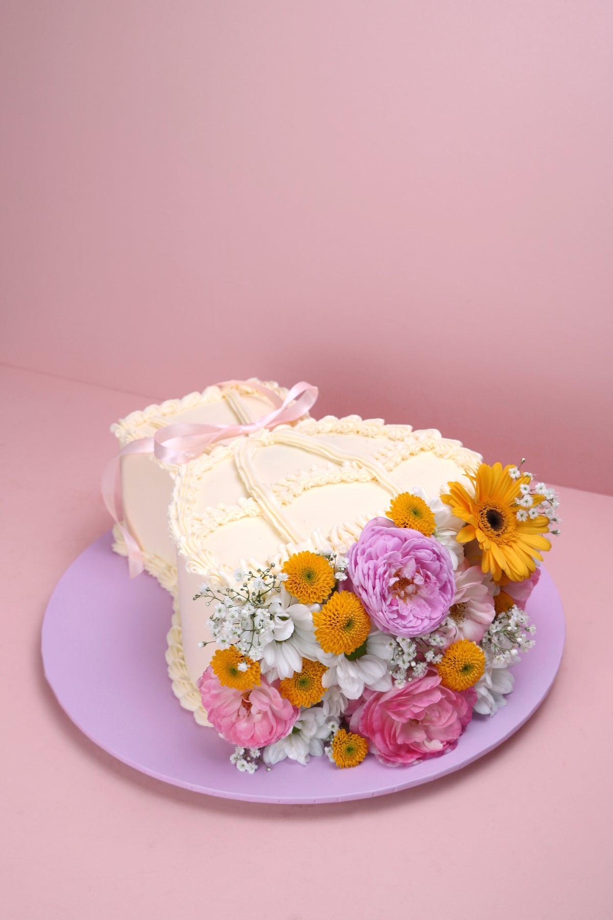 Heart-shaped fresh flower cake with yellow daisies, pink roses and vintage piped buttercream on lilac board handmade by Cakeasion in Uxbridge.