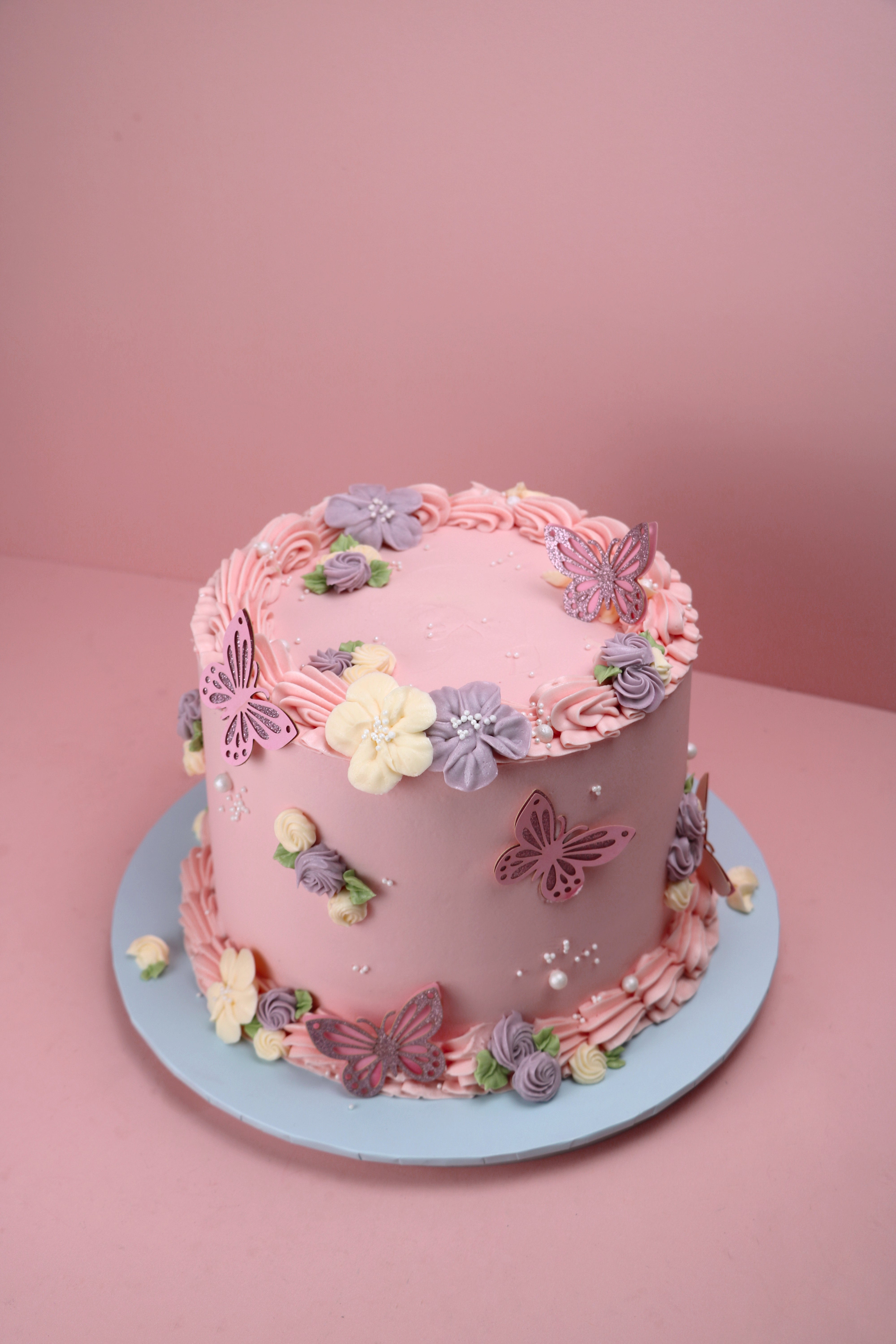 Pink round floral cake with butterfly toppers and piped lilac and ivory buttercream flowers – available in Cakeasion Uxbridge