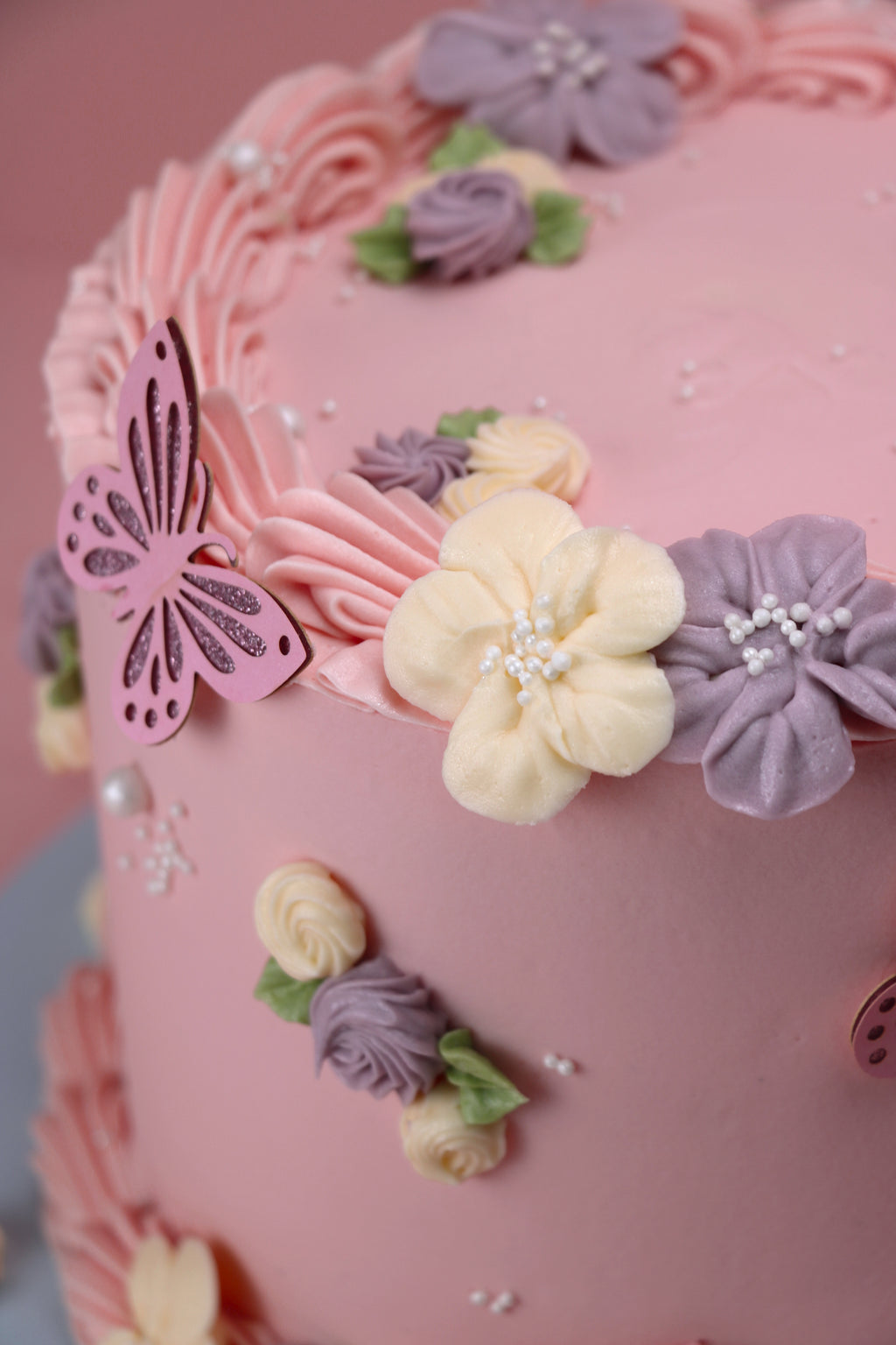 Close up of a Pink round floral cake with butterfly toppers and piped lilac and ivory buttercream flowers – available in Cakeasion Uxbridge